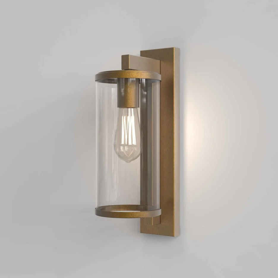 The Pimlico wall light is an outdoor light that features cylindrical glass against a rectangular backing in an antique brass finish.