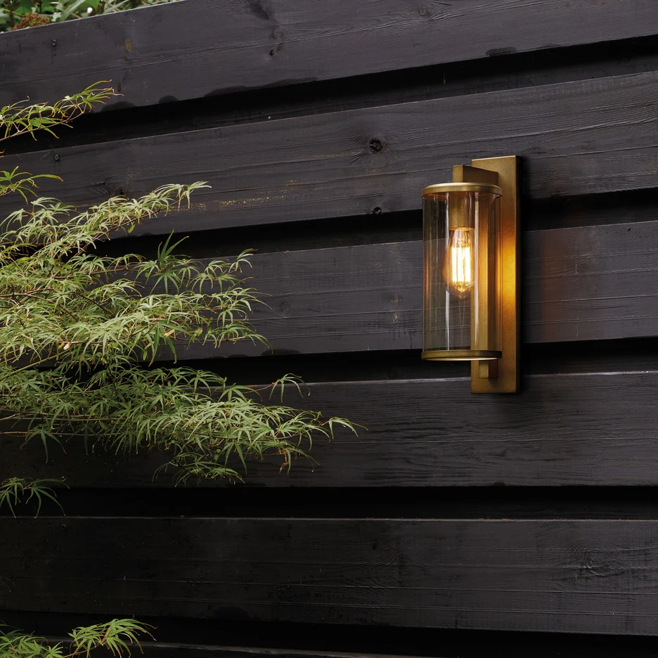 The Pimlico wall light is an outdoor light that features cylindrical glass against a rectangular backing in an antique brass finish. Shows the fitting installed outdoors.