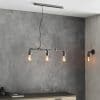 The Pipe pendant light has an authentic industrial design with 3 lamps on exposed pipe with aged pewter finish. Shows the light in a room hanging over a table with a grey backdrop. The matching Pipe wall light is featured on the opposing wall.
