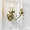 Shows the Bernice wall light on a plain white wall. The light features two curved arms finished in antique brass, supporting two traditional lampholders on either side. A pull cord switch hangs in the centre.