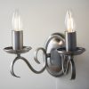 Shows the Bernice wall light on a plain white wall. The light features two curved arms finished in antique silver, supporting two traditional lampholders on either side. A pull cord switch hangs in the centre.