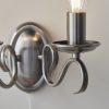 Close up of the Bernice wall light on a plain white wall. The light features two curved arms finished in antique silver, supporting two traditional lampholders on either side. A pull cord switch hangs in the centre.