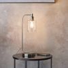 Shows the Toledo table light's lighting effect on a stand against a backdrop. The light features a classic design with a brushed nickel finish and cylindrical clear glass shade.