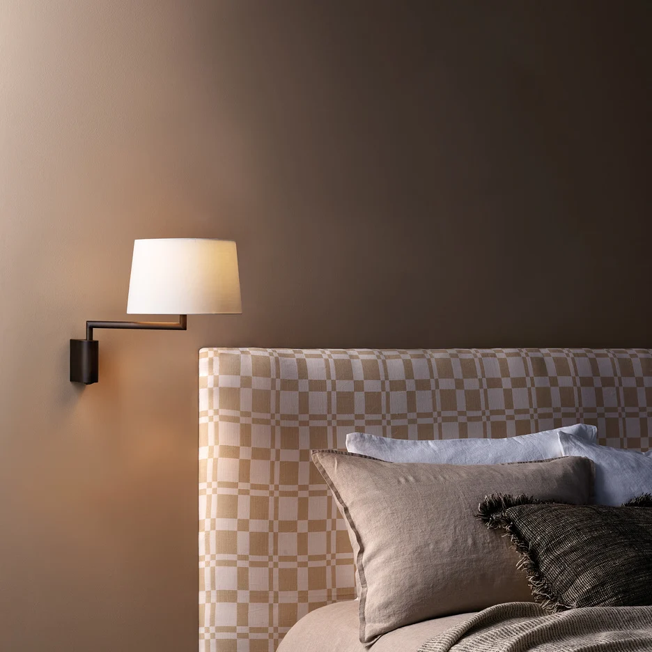 The Telegraph Swing wall light features a sleek, modern design with an angled arm and adjustable positioning in a bronze finish. Available with a tapered shade in four colours: white, black, putty, and mocha. Shows the fitting next to a bedside as a reading lamp.